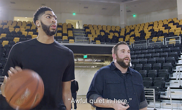 A man in a black shirt holds a basketball while another man stands beside him in an empty arena with yellow seats.