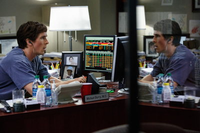 A man in a blue shirt sits at a desk with computer monitors, reflected in a glass partition.