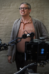 A man with glasses and a tan polo shirt under a grey jacket stands before a brick wall.