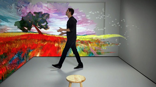 A man walks through a room where colorful, abstract landscape projections cover the walls.
