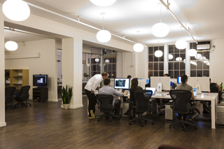 People sitting at desks with computer monitors in a brightly lit, modern open-plan office space.