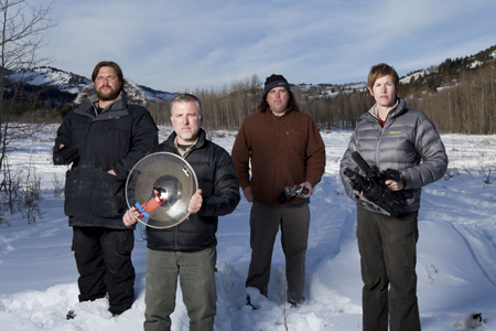 Four men stand in a snowy field with mountains and trees in the background, holding cameras and lighting gear.