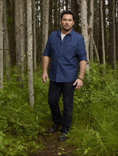 A man in a blue button-down shirt and dark pants standing on a path in a dense forest.