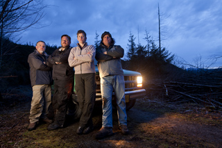Four men standing in a group in front of a vehicle in a wooded area at dusk.