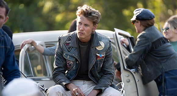 A man with blond hair wearing a distressed leather jacket sits on a vintage car outdoors.