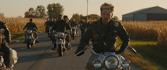 A group of people riding motorcycles down a paved road through a field at sunset.