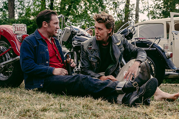 Two men sit on grass near motorcycles and a vintage car, one in a denim jacket and one in a leather jacket.