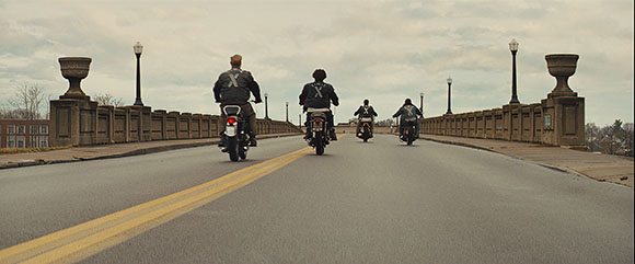 Four people riding motorcycles down the center of a wide bridge under a cloudy sky.