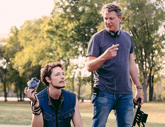 A man in a denim vest holds a camera while another man in a grey t-shirt gestures beside him.
