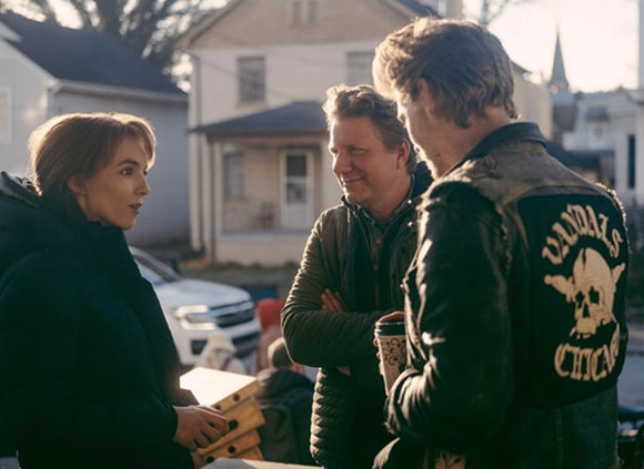 Three people stand together talking outdoors in front of a house during the day.
