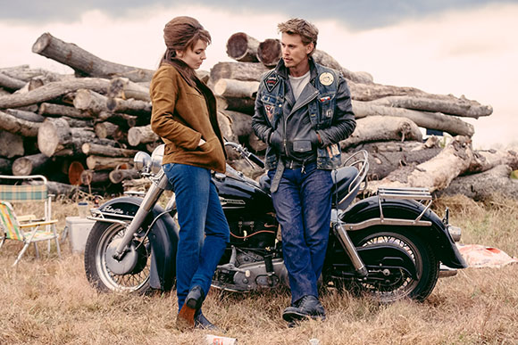 A man in a denim jacket and a woman in a brown jacket lean against a black motorcycle near large logs.