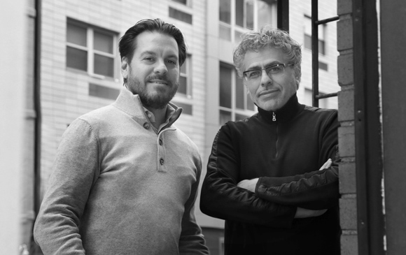 Two men posing outdoors in front of a multi-story building in black and white.