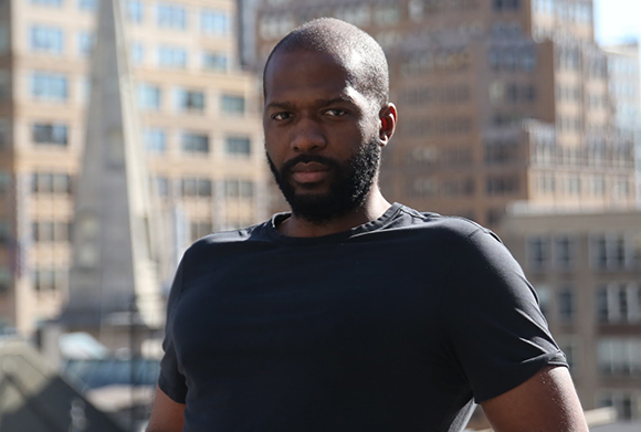 A Black man with a beard wearing a black t-shirt, looking at the camera against a blurred city backdrop.