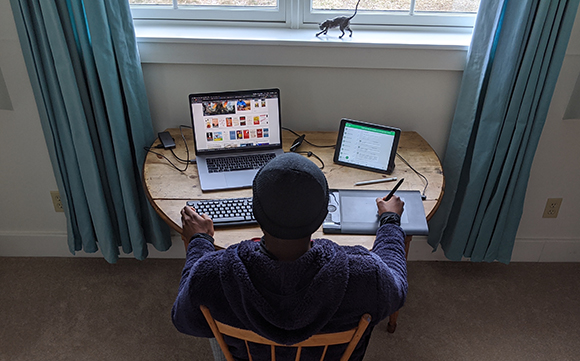 Overhead view of a person sitting at a wooden desk using a laptop, tablet, and digital drawing tablet.