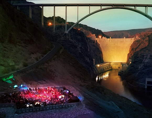 A large concrete dam and arch bridge span a dark canyon at dusk, with a crowd gathered at a lit event below.