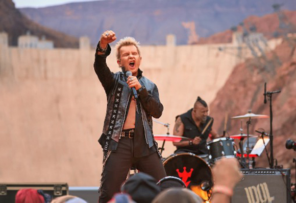 A male singer in a black leather jacket sings into a microphone on an outdoor stage.
