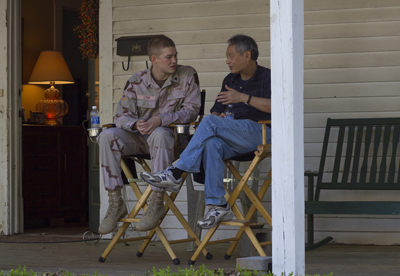 A man in military fatigues and a man in a dark shirt and jeans sit in director's chairs on a porch.