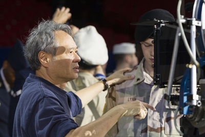 A man in a blue shirt gestures toward a camera while talking to an actor in military uniform.