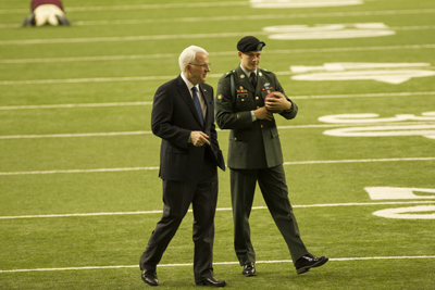 A man in a dark suit and a man in a green military uniform walk across a green football field.