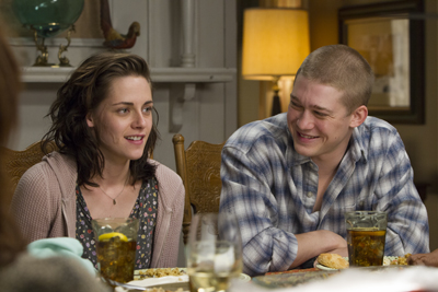 A woman with dark hair and a man in a plaid shirt smile at each other while sitting at a dinner table.