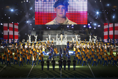 A marching band in yellow and blue uniforms performs on a field under a large screen showing a soldier.