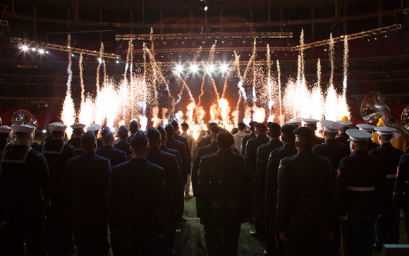 Service members in uniform stand in a stadium watching bright white fireworks erupt in the distance.