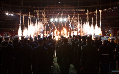 Military personnel in dark uniforms stand in a crowd watching white pyrotechnic sparks erupt in a large arena.