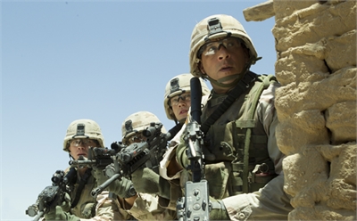 Four soldiers in combat gear and helmets hold rifles behind a textured mud wall under a clear sky.