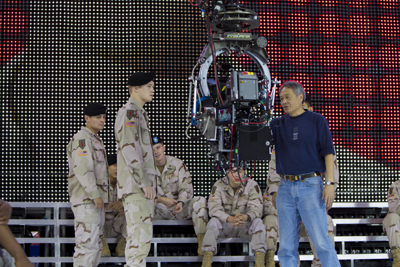 A film crew member stands near a large camera rig and soldiers in desert camouflage in front of an LED screen.
