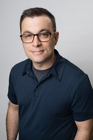 A man with short dark hair and glasses wearing a navy blue polo shirt against a light gray background.
