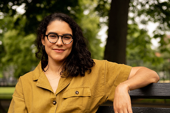 A smiling woman with dark curly hair and glasses wearing a mustard yellow shirt, leaning on a bench.