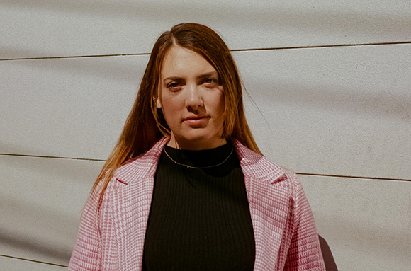 A woman with long brown hair wearing a pink blazer and black turtleneck against a textured wall.
