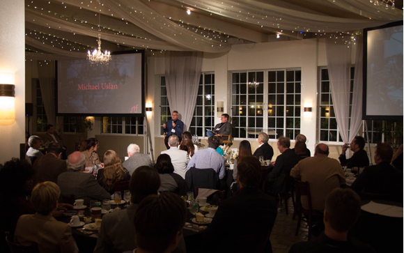 People seated at tables in a decorated room watching a presentation on a large screen.