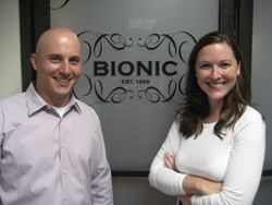 A smiling man in a light shirt and a woman in a white shirt standing before a Bionic company sign.