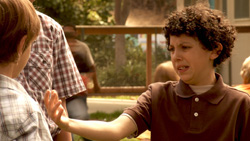 A boy with curly hair in a brown polo shirt gestures with his hand toward a person in a plaid shirt.