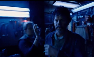 A man with dark hair and a beard looks toward the camera in a dark, blue-lit workshop.