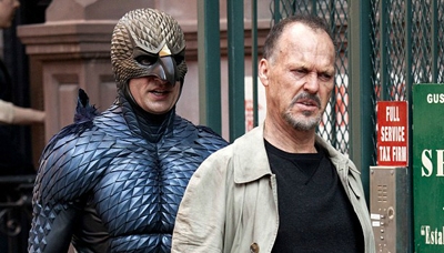 A man in a dark, scaled bird costume stands behind a man wearing a tan jacket and black shirt.