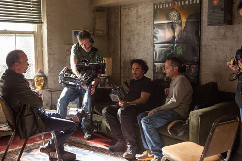A camera operator films two men sitting on a couch in a dimly lit room.