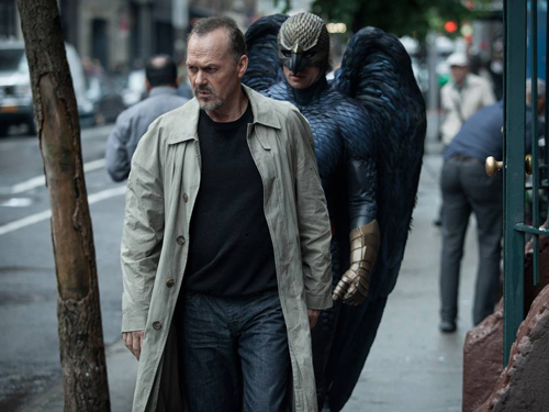 A man in a tan trench coat walks on a city sidewalk with a person in a dark feathered bird costume behind him.