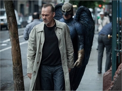 A man in a tan trench coat walks on a city sidewalk while a person in a dark bird costume follows closely.