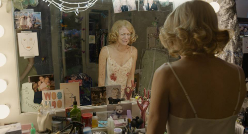 A woman in a white slip looks into a lit vanity mirror cluttered with photos and makeup.