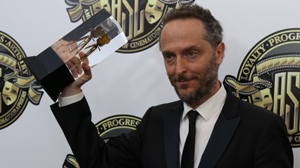 A man in a black suit and tie holds up a clear glass award trophy against a branded backdrop.