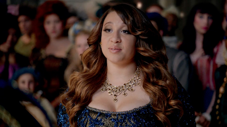 A woman with wavy brown hair wearing a blue sequined gown and a gold necklace stands before a crowd.