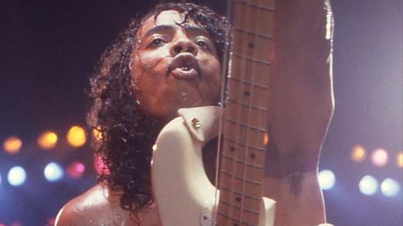 A person with long curly hair playing a white bass guitar on a dark stage with colorful lights.