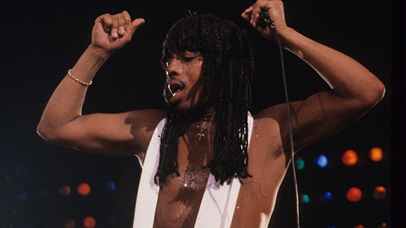 A person with long dreadlocks and a white vest singing into a microphone on stage.