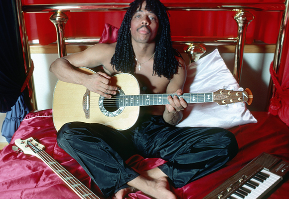 A man with long dreadlocks sits cross-legged on a red bed playing an acoustic guitar.