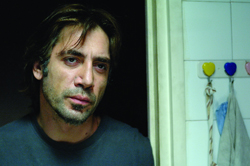 A man with dark, messy hair and facial stubble looking forward with a somber expression.