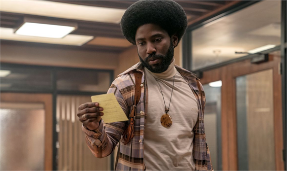 A man with an afro and a beard wearing a plaid shirt and turtleneck looks down at a small yellow slip of paper.