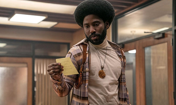 A man with a large afro and beard wearing a plaid shirt over a turtleneck looks down at a yellow slip of paper.