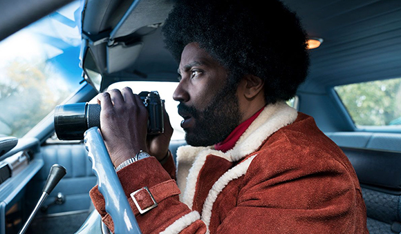 A man with an afro and beard wearing a shearling jacket looks through a camera lens while sitting in a car.